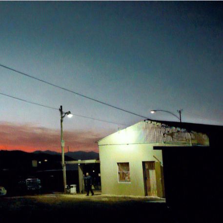 Seen from a distance, a lone figure walks from a dingy roadside restaurant under stark streetlight in the deepening twilight. Power lines stretch across the sky, which is deep blue at the top and red above the mountain range on the horizon.