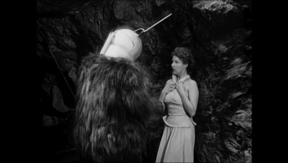 The robot monster confronting a woman who is clutching the straps of her dress tightly to herself