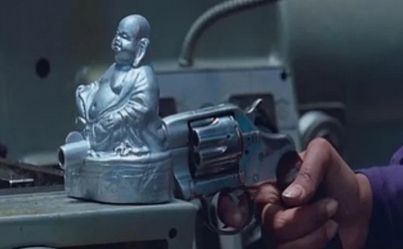 screencap of the buddha gun from the movie "the holy mountain". it's a silver statue of buddha built into the barrel of a revolver, held by a dark skinnedh and with a purple cuff. in front of a silver industrial background.