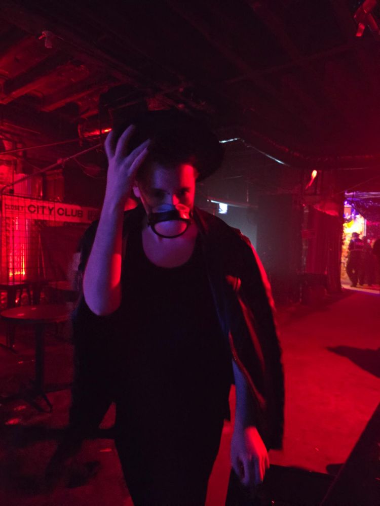 kinda blurry photo of Nic, a scrawny goth, in a low red-lit industrial club wearing a slutty little all-black ensemble. Their leather jacket draped over their bare shoulders, one delicate hand with black painted nails up on the brim of a black bolero hat. The only part of the outfit that isn't black is the white and gray of the ever-present Flo Mask