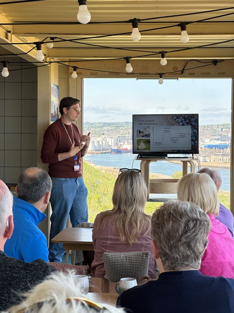 Me standing in the back, explaining what can be seen on the monitor next to me. In front of me is a crowd of people seated, and behind me shows the Aberdeen harbour