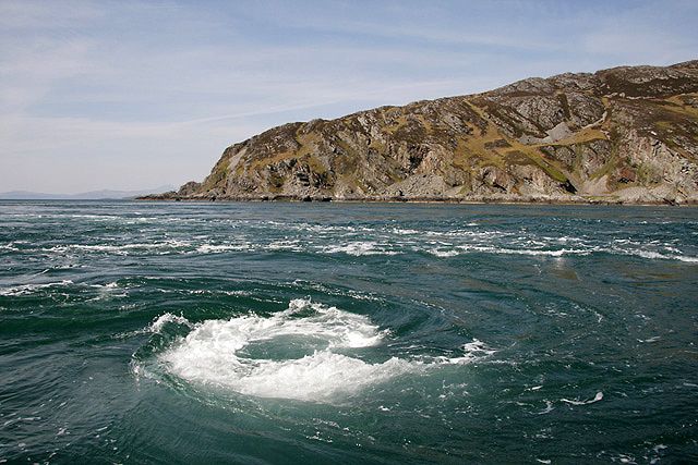 A picture of a large white spiralling whirlpool in the sea. The spiral shape is very distinct. In the background are hills that meet the sea. 