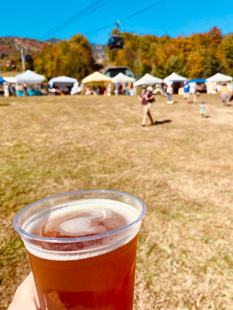 Beer with a festival in the background, with vendors under tents, a ski gondola overhead and a mountain in the background