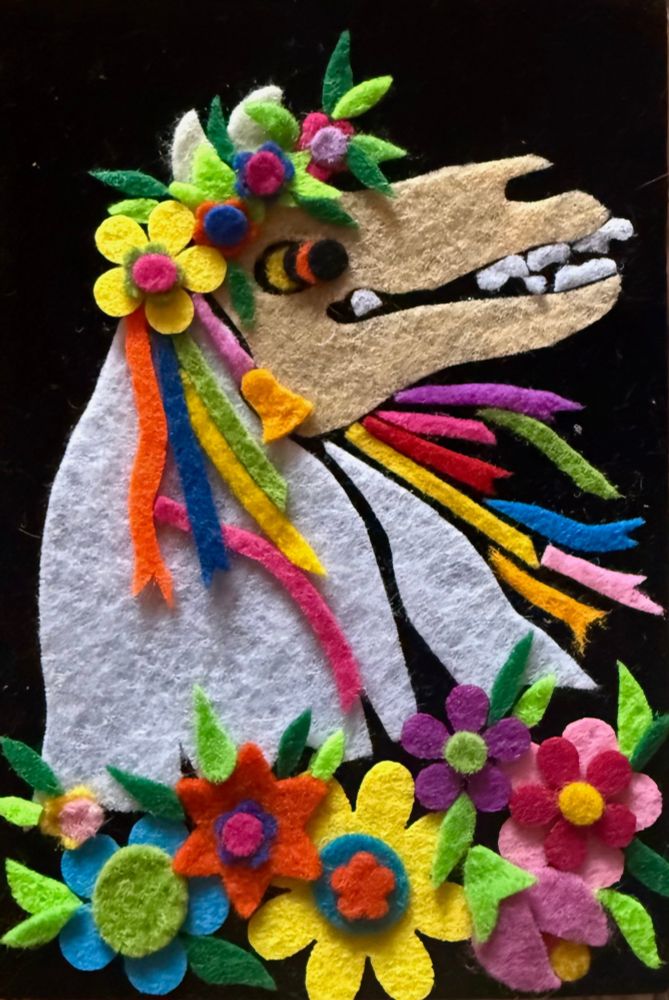 A brightly coloured Mari Lwyd surrounded by flowers all made of fuzzy felt on a black velvet background, the horse skull in profile facing right with their white sheet and rainbow ribbons billowing as they dance 