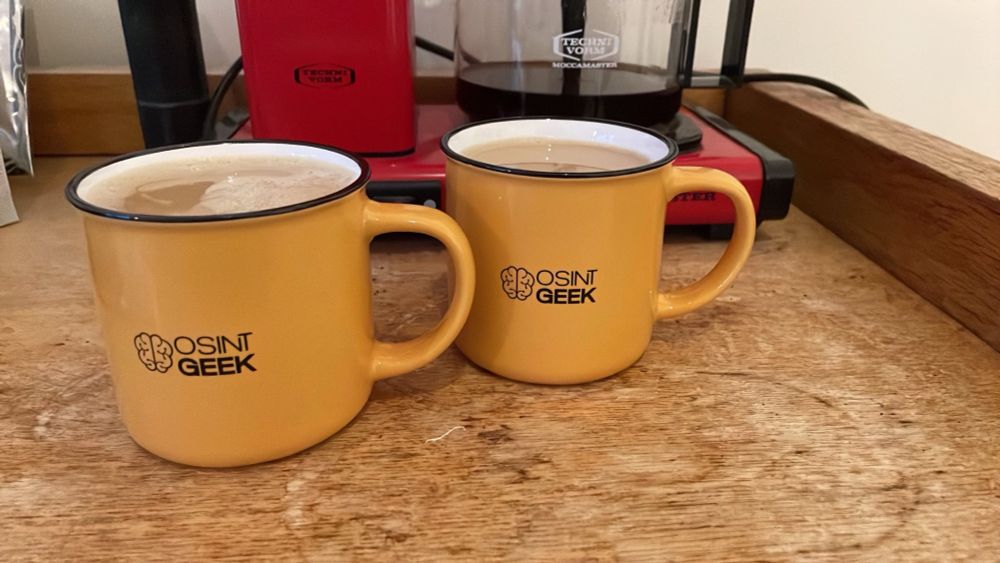 Zwei gelbe Tassen mit schwarzem OSINTgeek Logo stehen auf einem Tisch vor einer Kaffeemaschine.