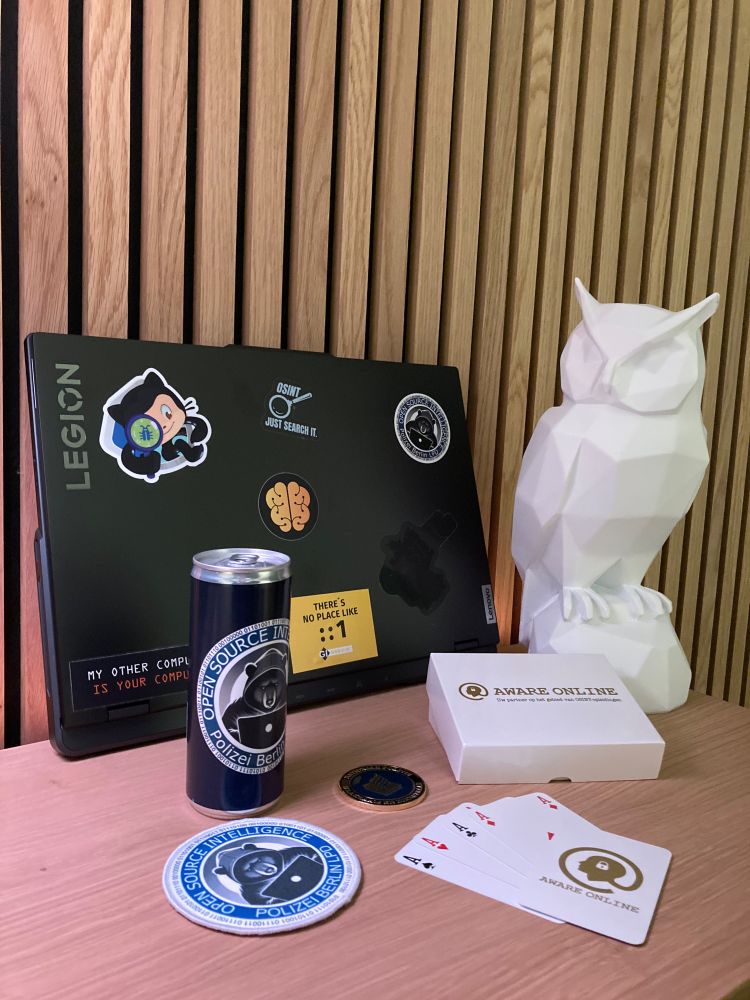 Ein stilvoll arrangierter OSINT-Setup auf einem Holztisch: Ein Lenovo Legion-Laptop mit mehreren OSINT-bezogenen Stickern, darunter ein GitHub-Octocat mit OSINT-Elementen, ein ‚Just Search It‘-Sticker und ein ‚There’s No Place Like :1‘-Sticker. Daneben eine weiße, geometrische Eulenfigur, eine Dose mit dem Logo der ‚Open Source Intelligence Polizei Berlin‘, ein passendes rundes Stoffabzeichen, ein Pokerkarten-Set. Außerdem liegt eine Challenge Coin auf dem Tisch.