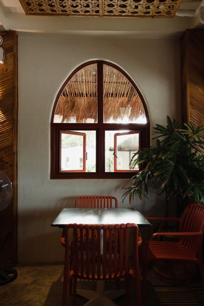 Photo of chairs placed around a square table, a leafy plant as decor on the corner on the right, a semi oval window with an overexposed view of the outside with nipa huts