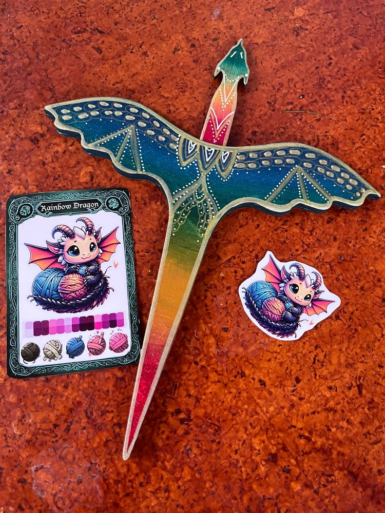 A rainbow painted plying dragon. Beside it are a card and sticker both featuring a baby dragon nesting in yarn. 
