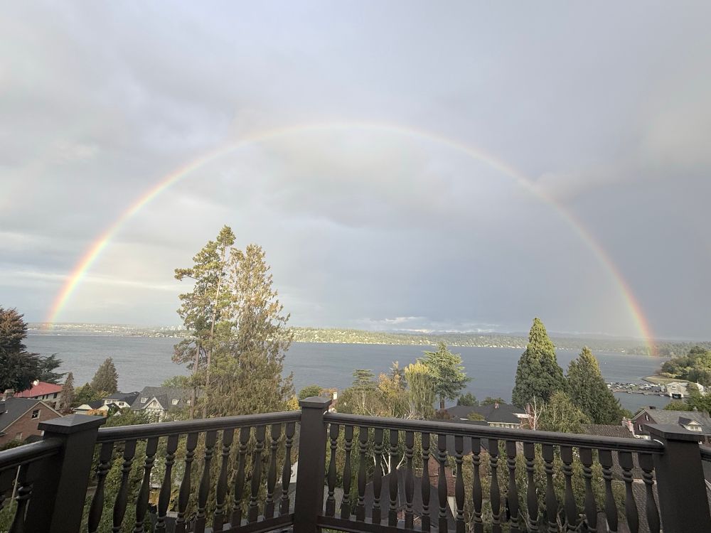 A perfect rainbow - viewed from a deck w brown railing - over the lake w trees in the foreground. 