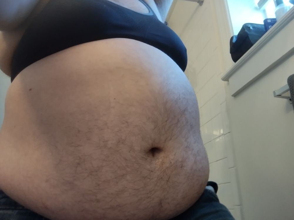 Transgender man wearing a black binder showing off his large hairy belly, wearing jeans. 