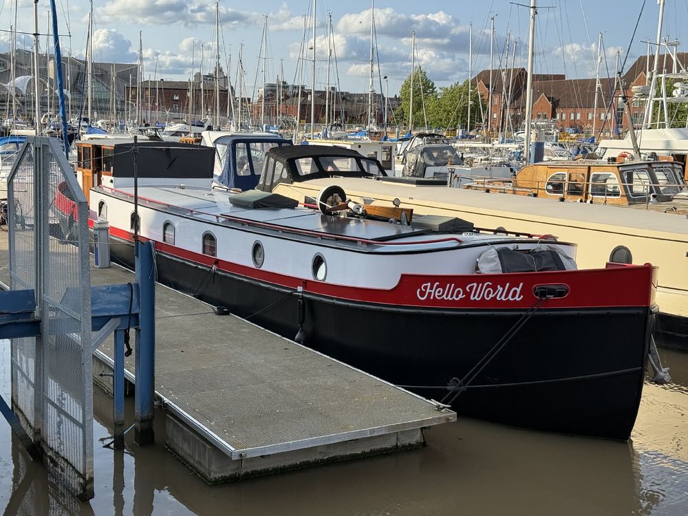 A black and red boat named "Hello World" docked at a marina with other boats. The boat has a white cabin and a red stripe along the side. The marina is filled with various boats and has a blue dock. The sky is blue with some clouds. There are buildings and trees in the background. A metal ramp is leading up to the boat. The water is calm and reflects the boats and the sky.