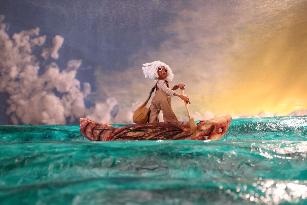 A stop-motion puppet on a set. The puppet is standing up in a canoe-type boat carved like a red dragon, and she’s holding an oar. She is turned to look over her shoulder at the camera. She has brown skin, white short hair, brown overalls and a checkered shirt. She is traveling across turquoise waters, with a yellow sunset beginning on a blue sky spotted with clouds.