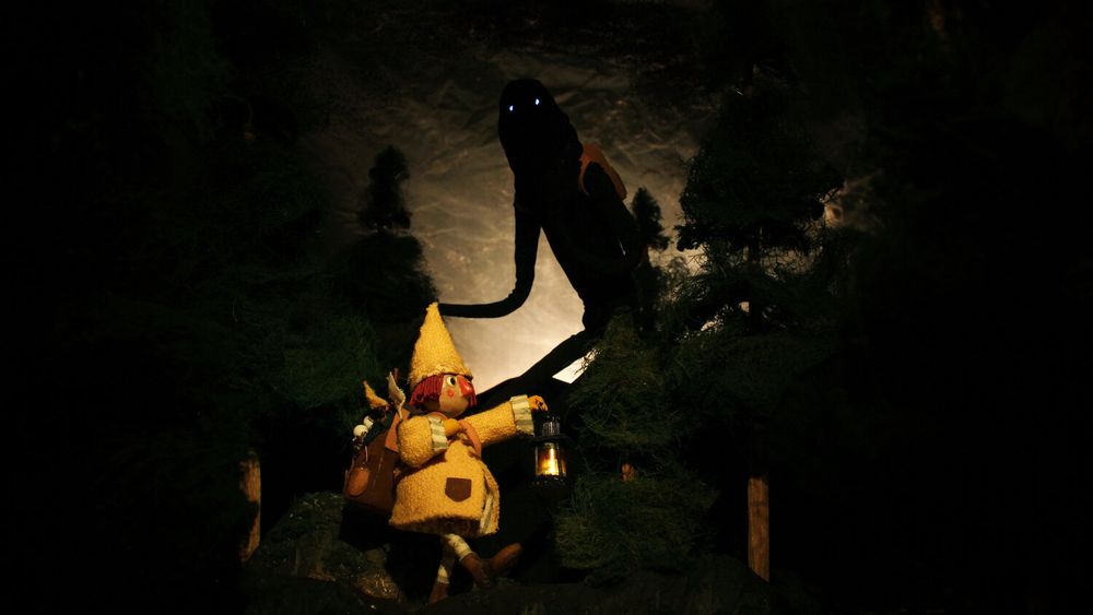 Stop motion puppets photographed on a set. The first is lit by a patient hey are holding, and they are marching to the right. They have a big yellow wool coat, a yellow pointy hat, short magenta hair, and an overstuffed backpack. They are surrounded by dark pine woods. Behind them, silhouetted, is a giant shadow, with just two pinpricks of light for eyes. They are wearing a little orange backpack. The sky is lit by a low moonlight.