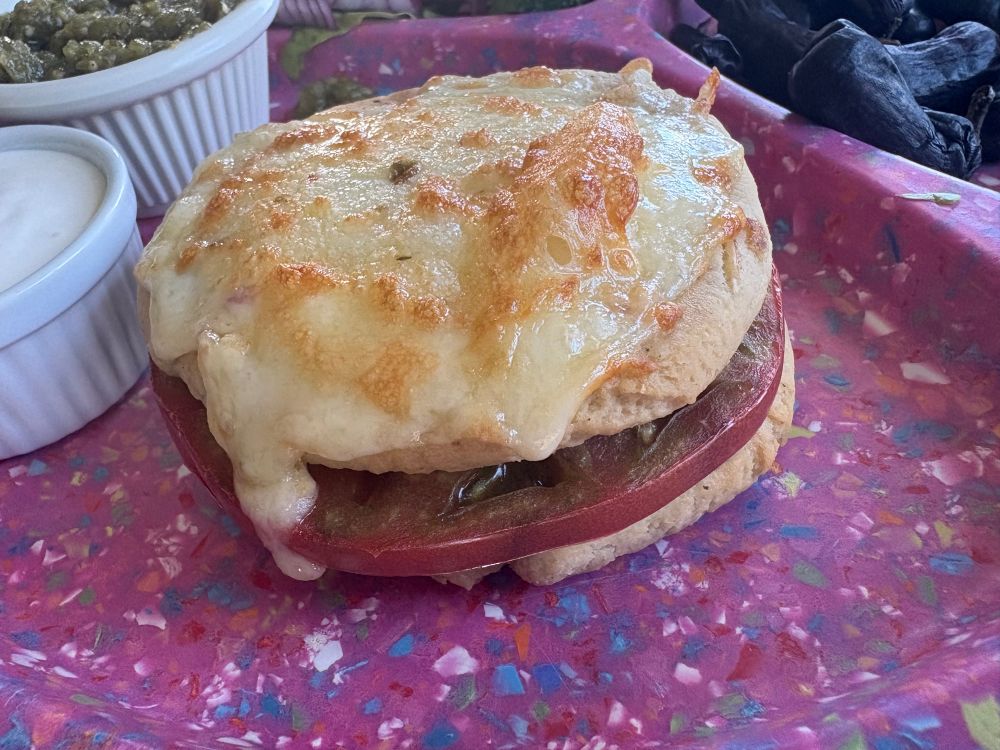 A slice of tomato between two papusas, topped with browned cheese 