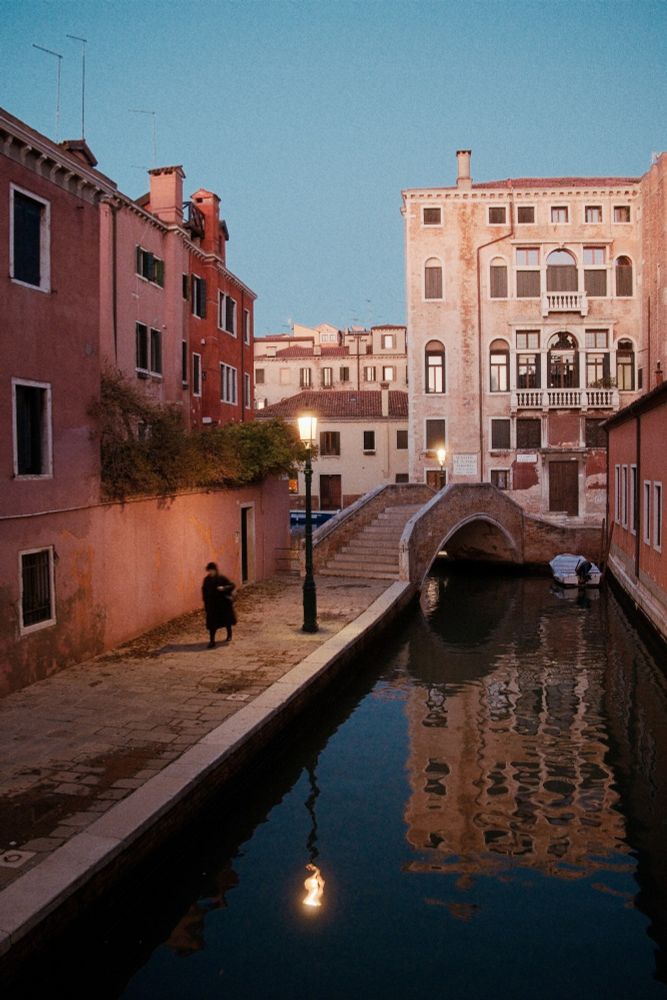 An image of Venice at twilight. A woman crosses the bridge over a canal, under the glow of a street lamp.
