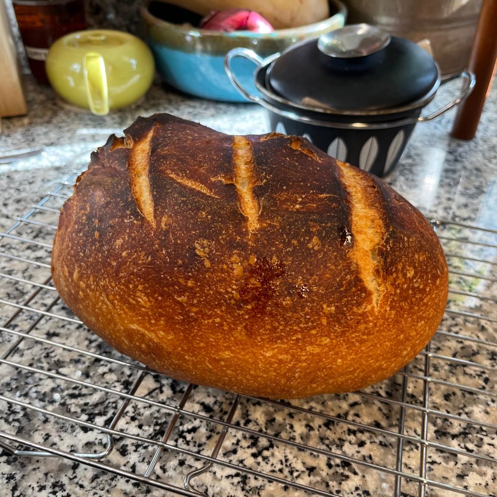 Loaf of baked sourdough bread sitting on wire rack on granite countertop.