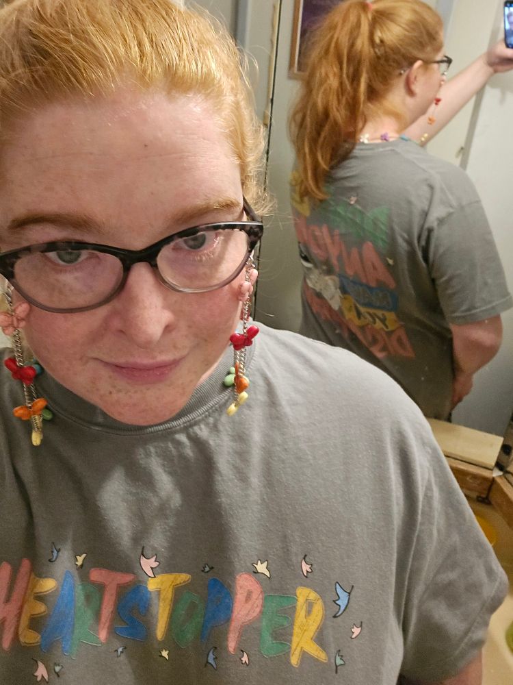 Selfie in front of mirror of a lady type person with red hair, glasses with a colorful glasses chain, and a gray Heartstopper shirt