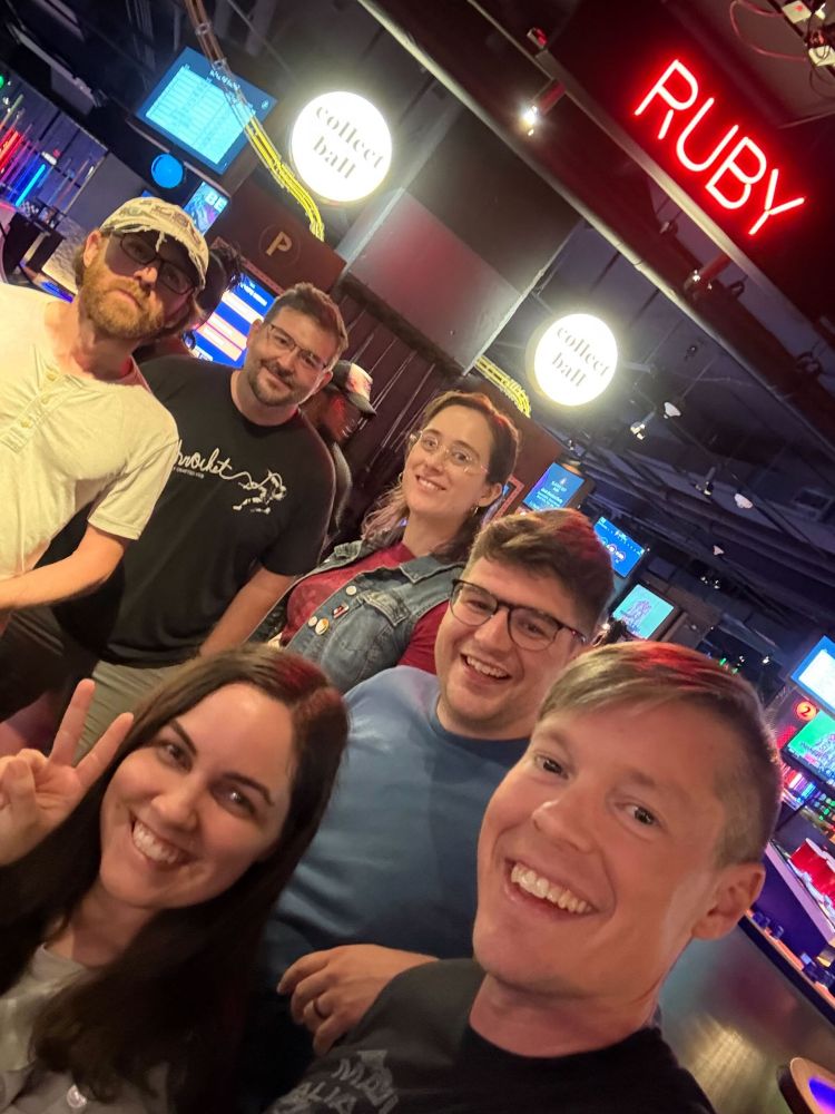 Thomas, Matt, Chael, Thomas, Kat, and Jeremy all smiling under a glowing neon Ruby sign.