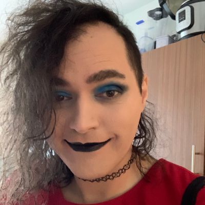 Vexx Daniel: A portrait of a brunette trans woman smiling, wearing makeup and a red dress, with a sidecut.