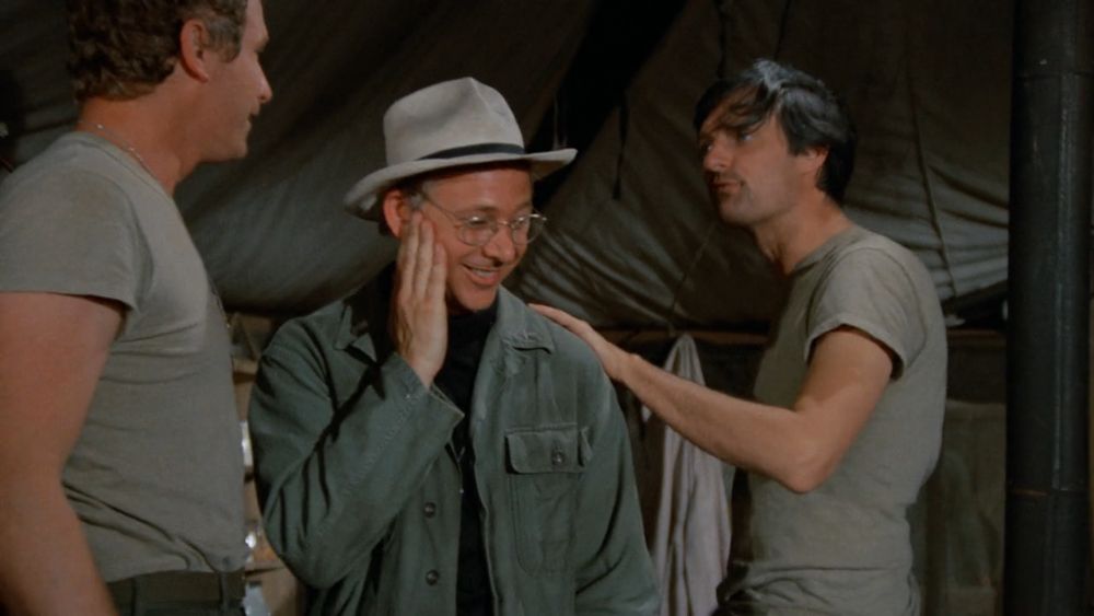 Mulcahy touches the spot on his cheek where Trapper kissed him, still grinning and looking a bit stunned, and Hawkeye looks mildly annoyed