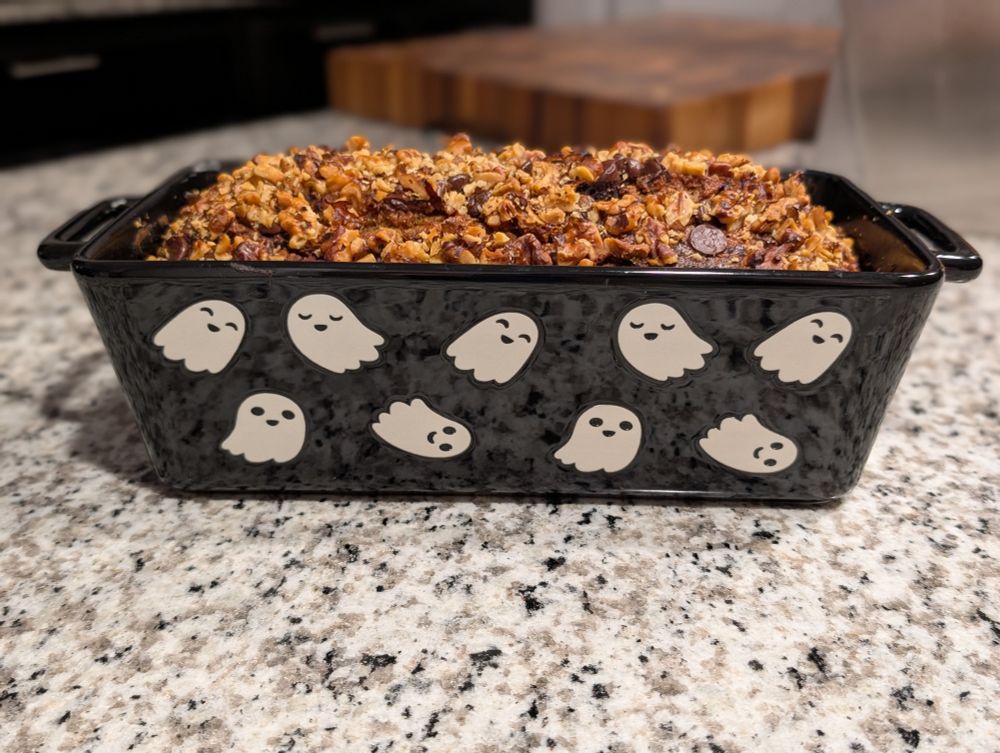 Side view of a loaf of chocolate banana bread topped with walnuts in a black ceramic baking dish patterned with white ghosts.