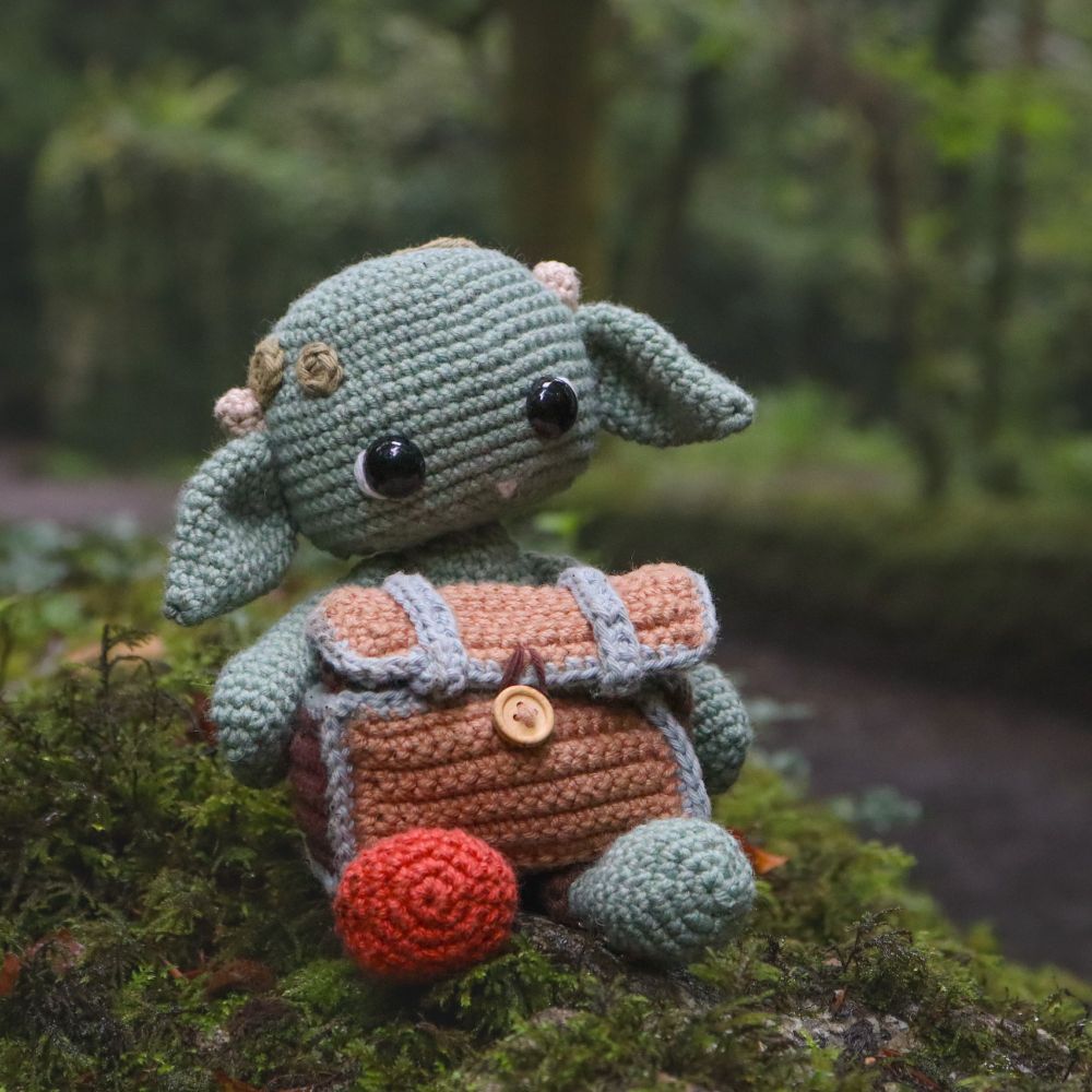 Image of a crocheted goblin who is holding a treasure chest. The goblin is a light sage green and has big ears and a very cute snaggle tooth. 

They are sitting on some moss in a green forest. 