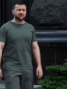a man with a beard wearing a green t-shirt and gray pants is standing in front of a building .