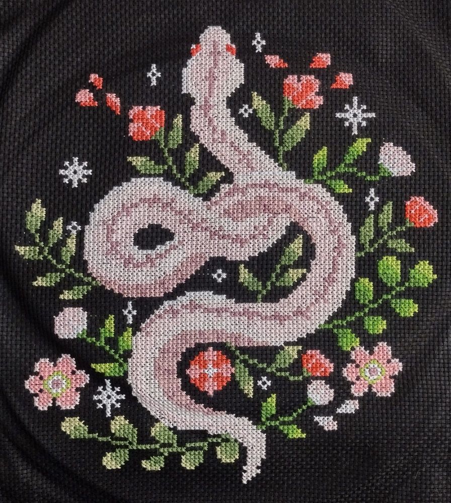 A cross stitch of a pink snake with flowers and stars on a black background.