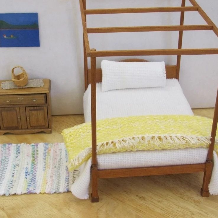 Handcrafted Inch to Foot Dollhouse Miniature Herringbone Bedspread, Handmade Bedding Throw Bed Blanket Artisan Hand Loom Woven in Sunny Yellow & White, 1:12 Scale Doll House Bedroom Home Decor Furniture Accent. Styled folded on the end of a bed with white bedspeard and pillow. A hand woven rag rug is on the floor, a small side cabinet with a handwoven linen runner and straw basket, and a modern waterscape painting is on the wall.