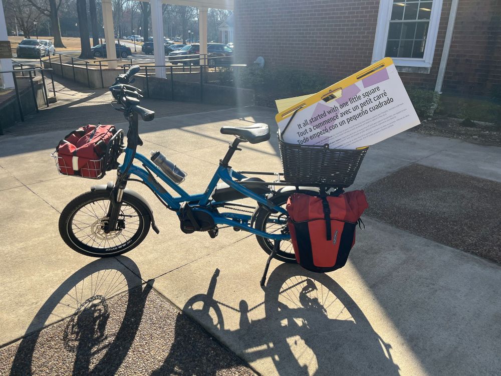 A cargo bike loaded down with a big red pannier and some large teaching supplies in a basket on the back rack.