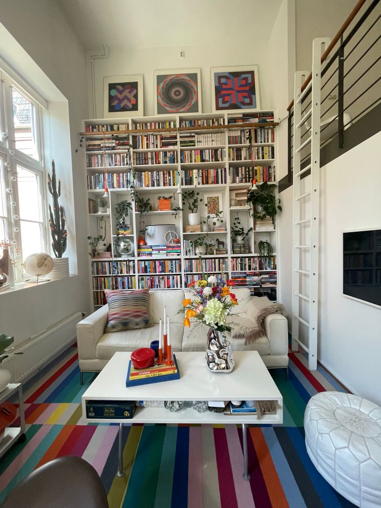 Bright white apartment with floor to ceiling shelves filled with a rainbow of books and plants. Striped rainbow rugs, white furniture, and a colorful bouquet of are also present. 