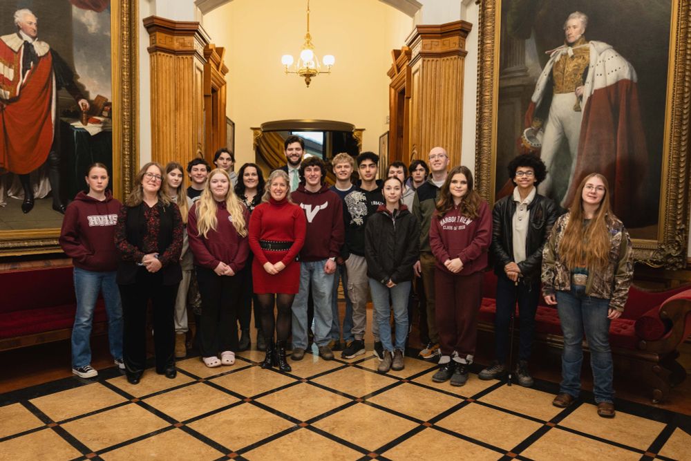 A group of people stands in a room with ornate decor, posing for a photo. They are surrounded by large portraits in an elegant hallway with patterned flooring and wooden columns. Some are wearing matching maroon shirts.
