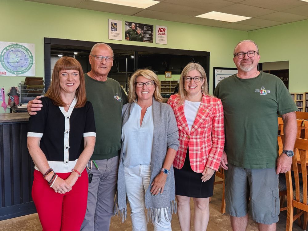  Five adults stand together indoors, smiling at the camera. Three people wear casual green shirts, while two women wear business attire—one in a red plaid blazer and another in a black and white top with red pants. -- Cinq adultes se tiennent ensemble à l'intérieur et sourient à l'appareil photo. Trois personnes portent des chemises vertes décontractées, tandis que deux femmes portent des vêtements d'affaires, l'une avec un blazer rouge à carreaux et l'autre avec un haut noir et blanc et un pantalon rouge.