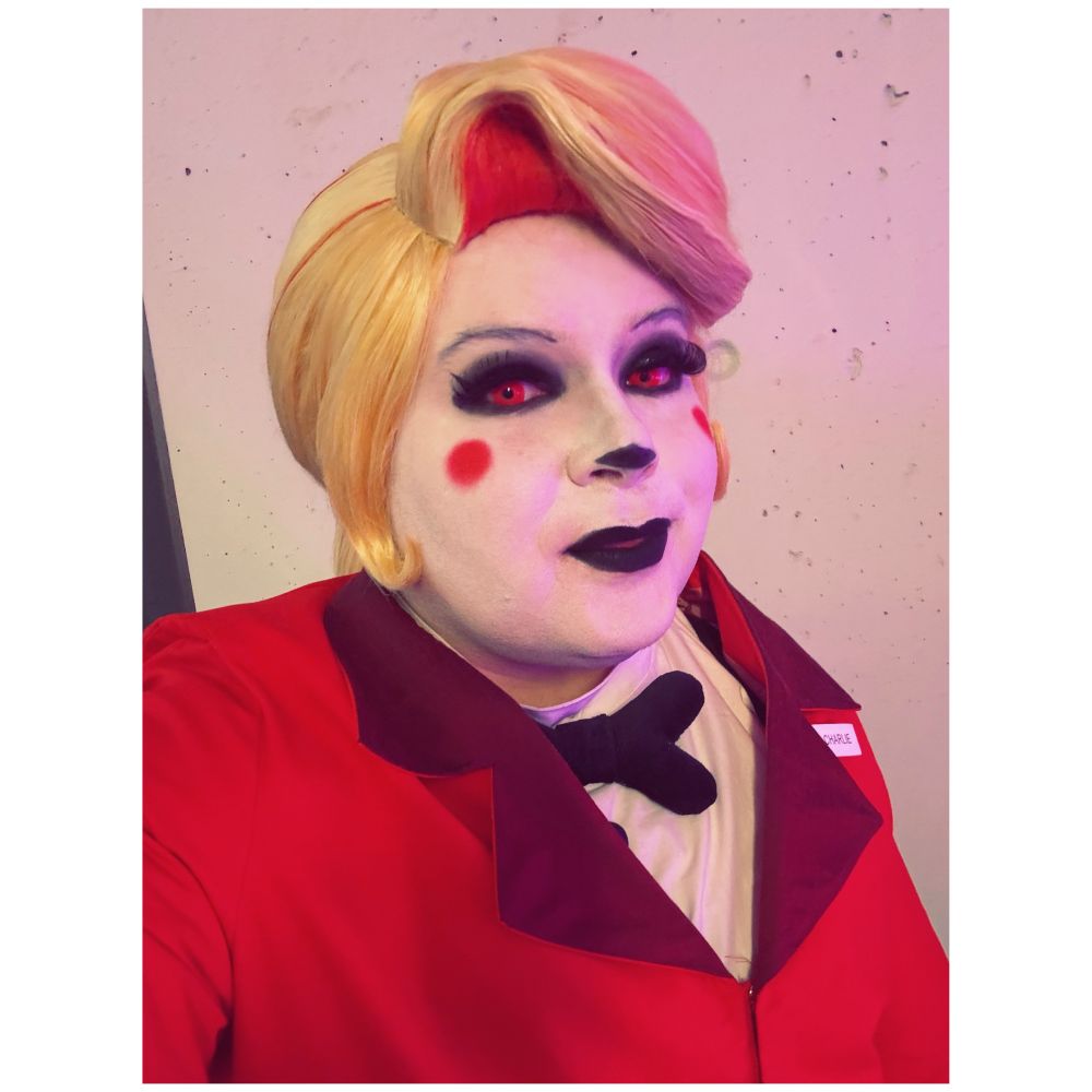 Selfie of Alex wearing a Charlie Morningstar (Hazbin Hotel) cosplay. It includes red pantsuit, black bow tie and a white shirt. The make up is white foundation, heavy black make up, red dots on the cheeks and black lipstick.
