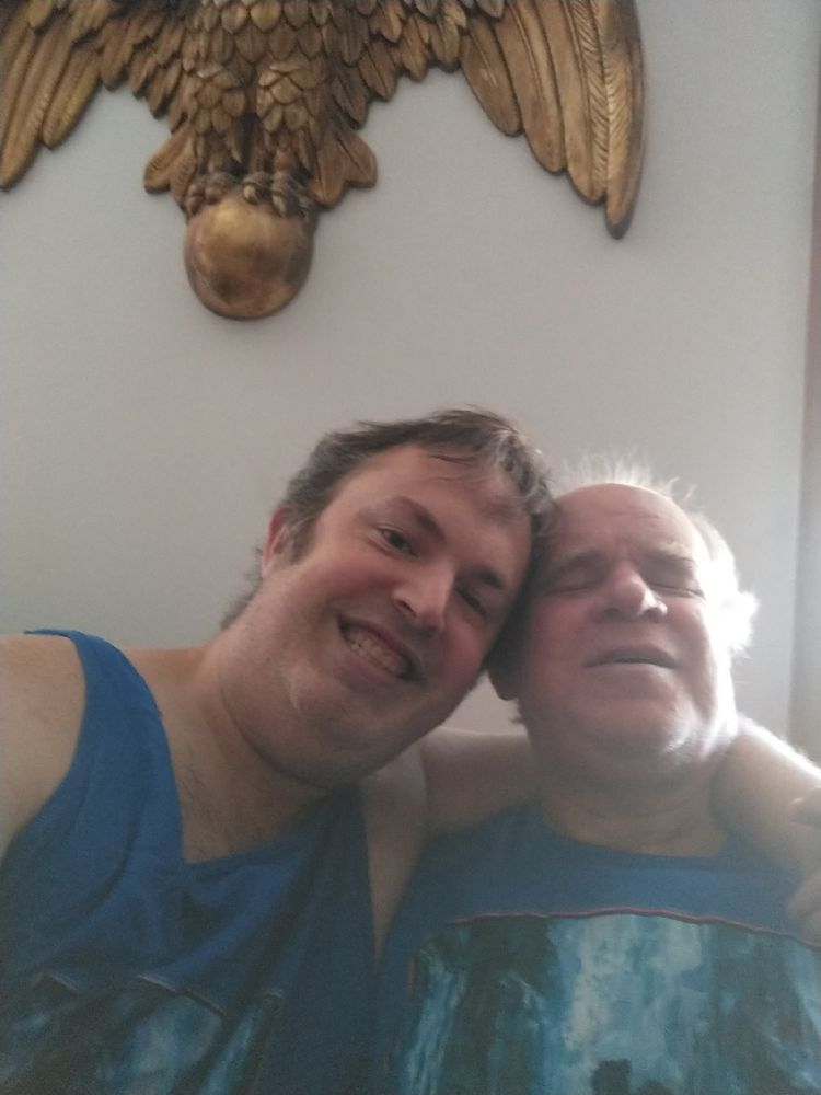 The photograph depicts two men in an indoor setting, posing for a picture. The man on the left is younger, with short hair and a beard, wearing a blue sleeveless shirt. He is smiling broadly, showing his teeth. The man on the right is older, with white hair and a white beard, also wearing a blue sleeveless shirt with a graphic design. He is leaning against the younger man, with his eyes closed, appearing relaxed or perhaps sleepy. Behind them, a large bronze-colored eagle head is mounted on the wall, adding a touch of grandeur to the otherwise simple setting. The wall is plain, with a neutral color, and there are no other visible decorations or objects in the immediate vicinity. The overall atmosphere is one of camaraderie and comfort between the two men.