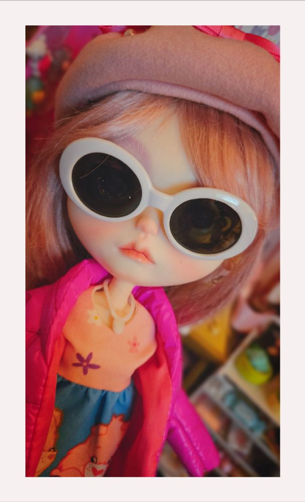 Close-up of my big-headed Blythe doll. She's wearing a pink wintery outfit, a pink hat, and white sunglasses. Her hair is purple-pink and she look like she has an attitude 