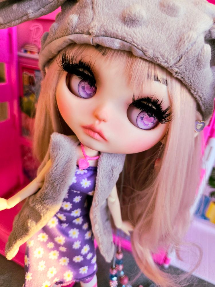 A photo of my large-headed doll with pale pink hair and pink heart eyes. She's wearing a gray bunny ear hat and looking to the right with a tilted head