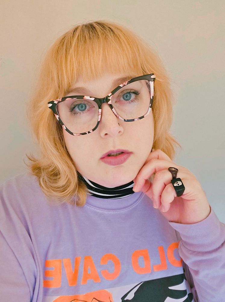 A photo of me. I have shoulder-length blonde hair and blue eyes. I'm wearing black glasses and a purple sweater. I have my hand on my chin