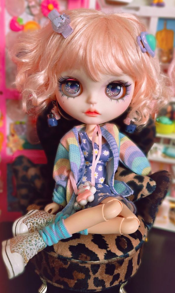 Big-headed pink-haired doll. She's wearing a colorful purple and green outfit and sitting casually in a big comfy chair, looking at camera