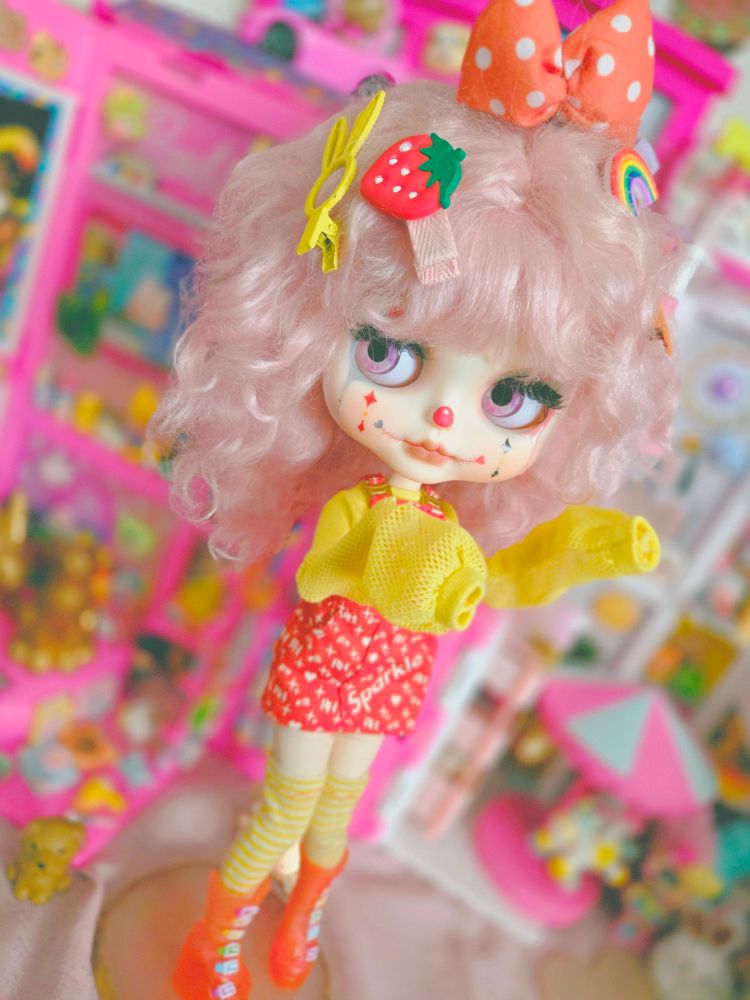 Big-headed clown doll with pink eyes, pink hair, and a colorful red and yellow outfit. She's looking left and her oversized sleeves are hanging over her hands