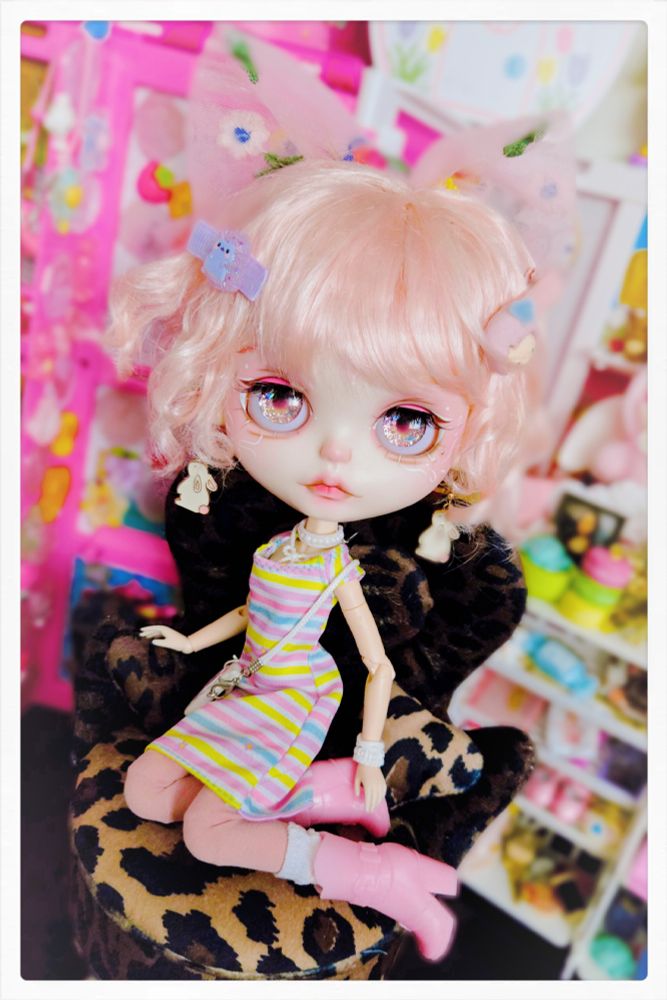 Pink-haired big-headed Blythe doll with golden sparkling eyes. She's wearing a colorful pink and yellow outfit with pink cowboy boots and bows and clips in her hair. She is seated on a leopard print chair with her legs folded underneath her