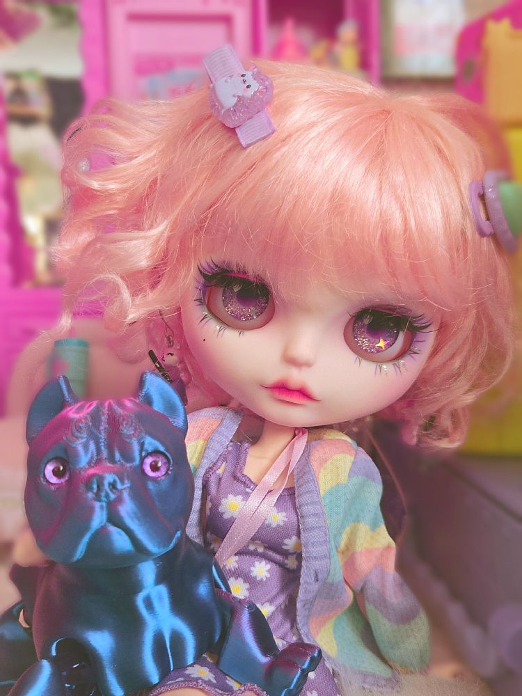 Big-headed Blythe doll with a short curly pink bob and sparkly purple eyes. She's sitting on the floor and holding her blue bulldog 