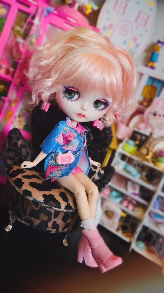 Big-headed Blythe doll sitting on a leopard print chair. She had a curly pink bob and is wearing a summery blue and pink outfit. Seated casually, she's looking to the left sleepily 