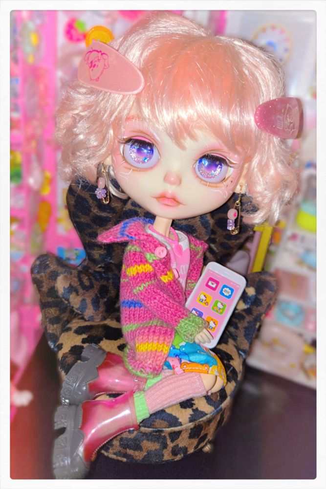 Pink-haired Blythe doll with a big head and a short curly bob. She's wearing a cozy pink outfit with a long sweater and big boots. She's sitting on a leopard print chair with a giant Hello Kitty cell phone