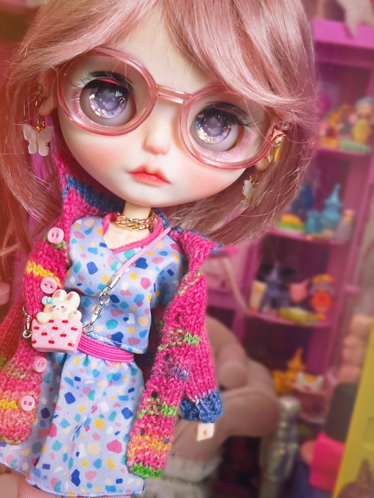 Big-headed Blythe doll with pink-purple hair. She's wearing pink glasses and a colorful pink and purple outfit. She's looking at the camera with purple eyes