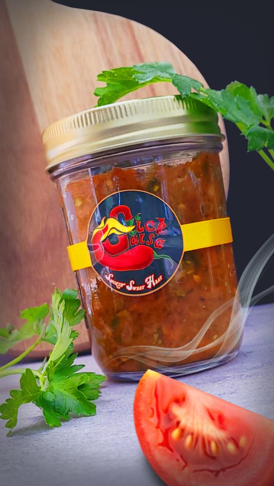 A jar of smoked salsa. Adorned on the jar is branding Slick Salsa, underneath it says Smokin' sweet heat. The product photography composition consists of a few sprigs of parsley, a tomato wedge, a wispy smoke trail, and a chopping board. A concrete, marble countertop and a darkened backdrop.