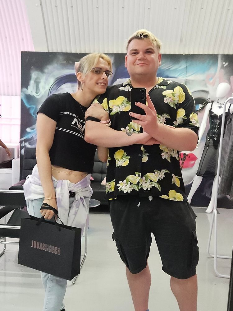 JB and Ashley in the mirror of charm school, Ashley is holding onto JBs bicep and holding a Charmskool shopping bag.