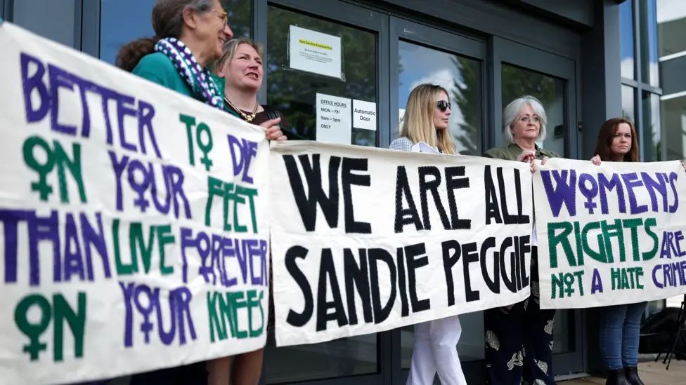 Bunch of terfs holding up a banner saying "we are all sandie peggie" 
