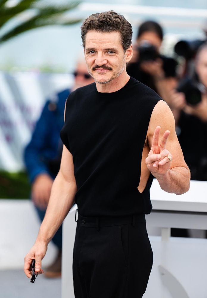 Pedro Pascal, in a sleeveless black shirt that is open almost down to his waist and fitted black slacks, flashing a peace sign.