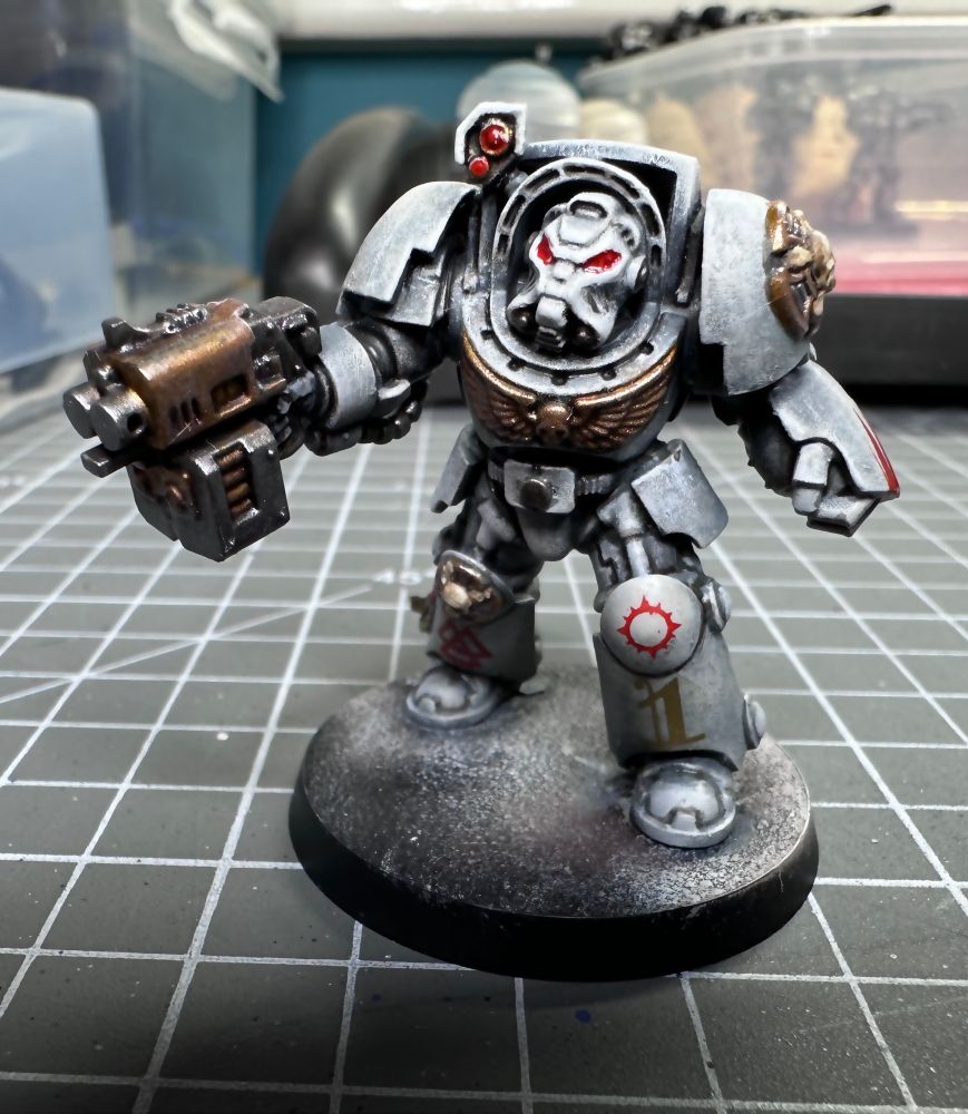 A photo of a terminator space marine wearing white power armour. His left hand is in a power fist and his right hand holds a storm bolter gun. The details are in gold with some red touches. 